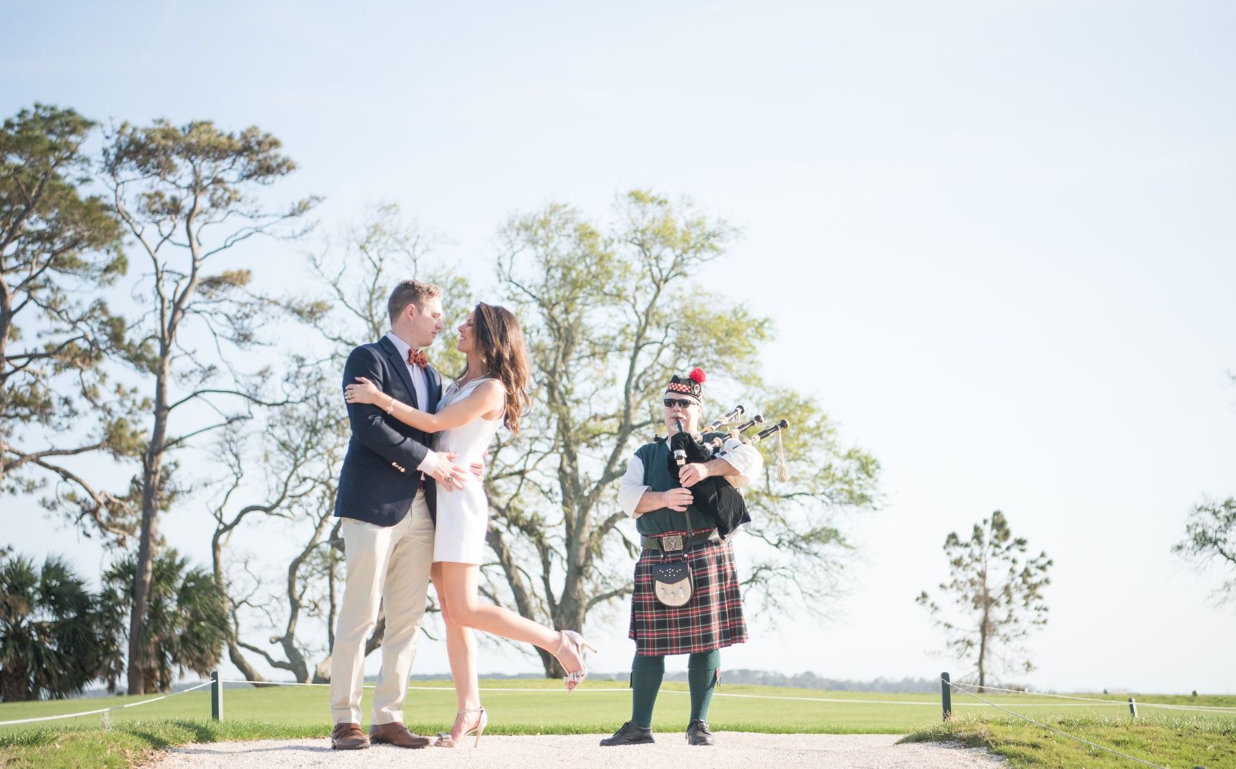 bride and groom hugging on a golf course with a man playing bagpipes behind them