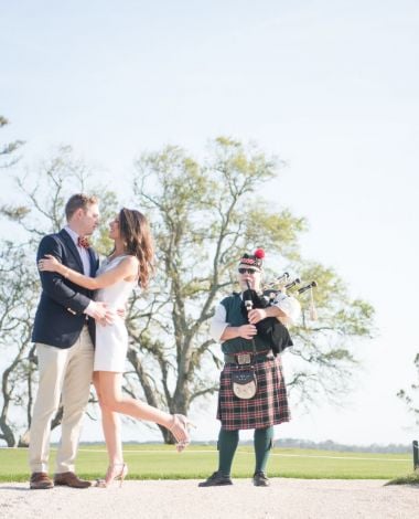 bride and groom hugging on a golf course with a man playing bagpipes behind them
