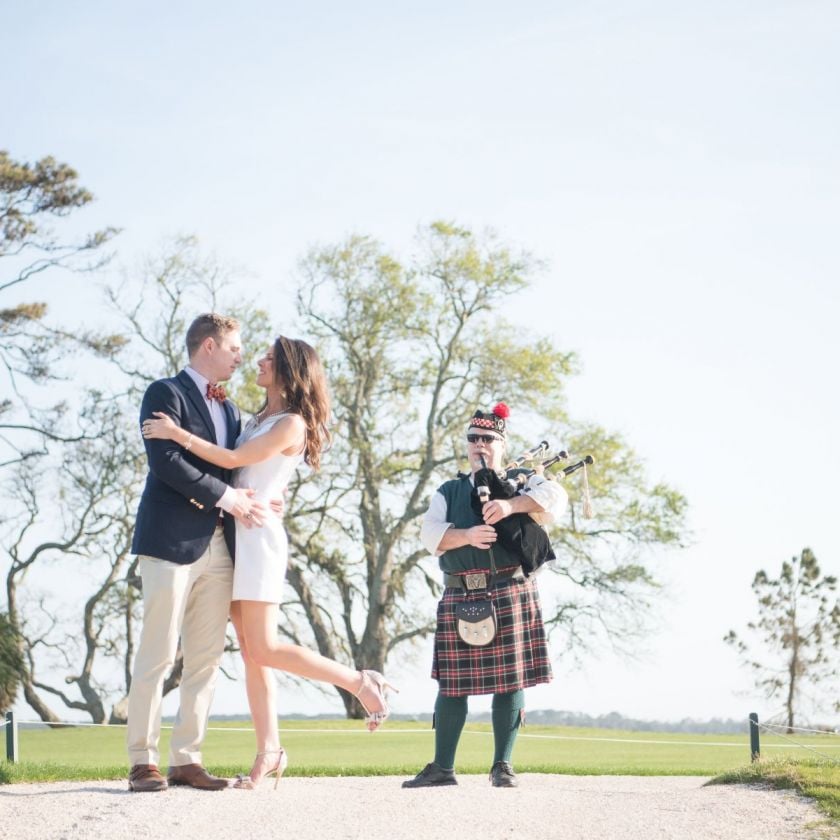 bride and groom hugging on a golf course with a man playing bagpipes behind them