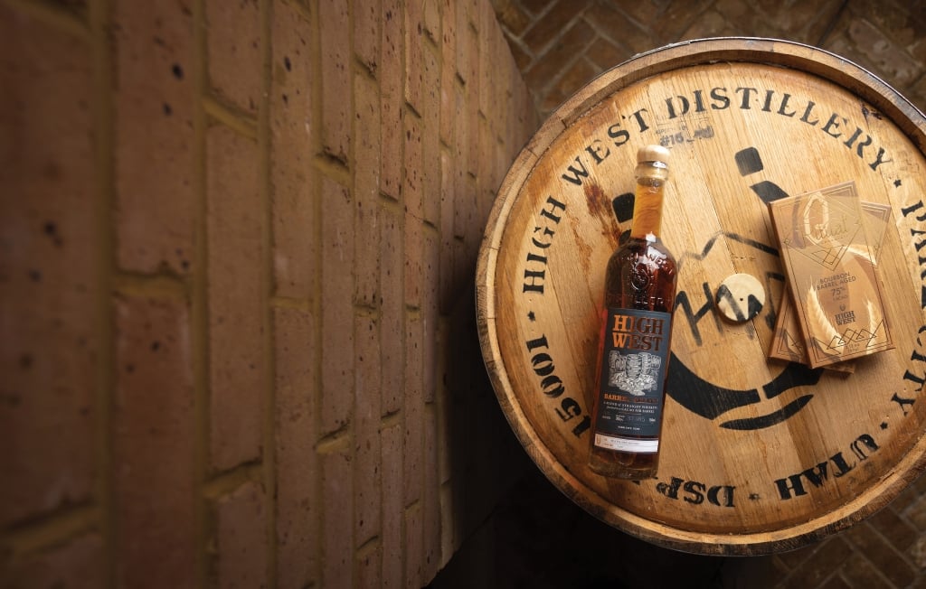 A bottle of high west, on a barrel next to a brick wall.