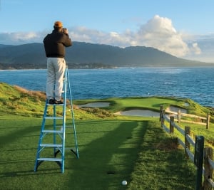 Evan Schiller photographing the iconic 7th hole at pebble beach golf links, from atop a ladder, on a beautiful December afternoon.