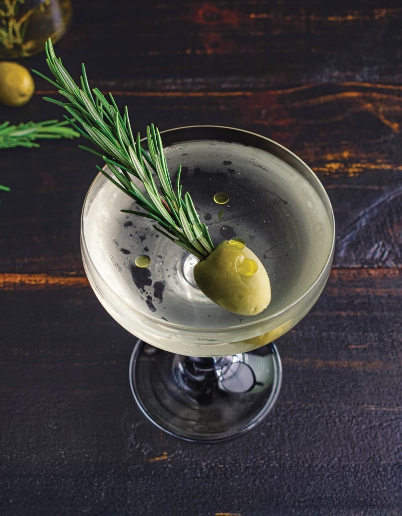 Smoked Rosemary Oil Dirty Martini: A dirty gin martini served up in a coupe glass and garnished with a sprig of rosemary in a green olive