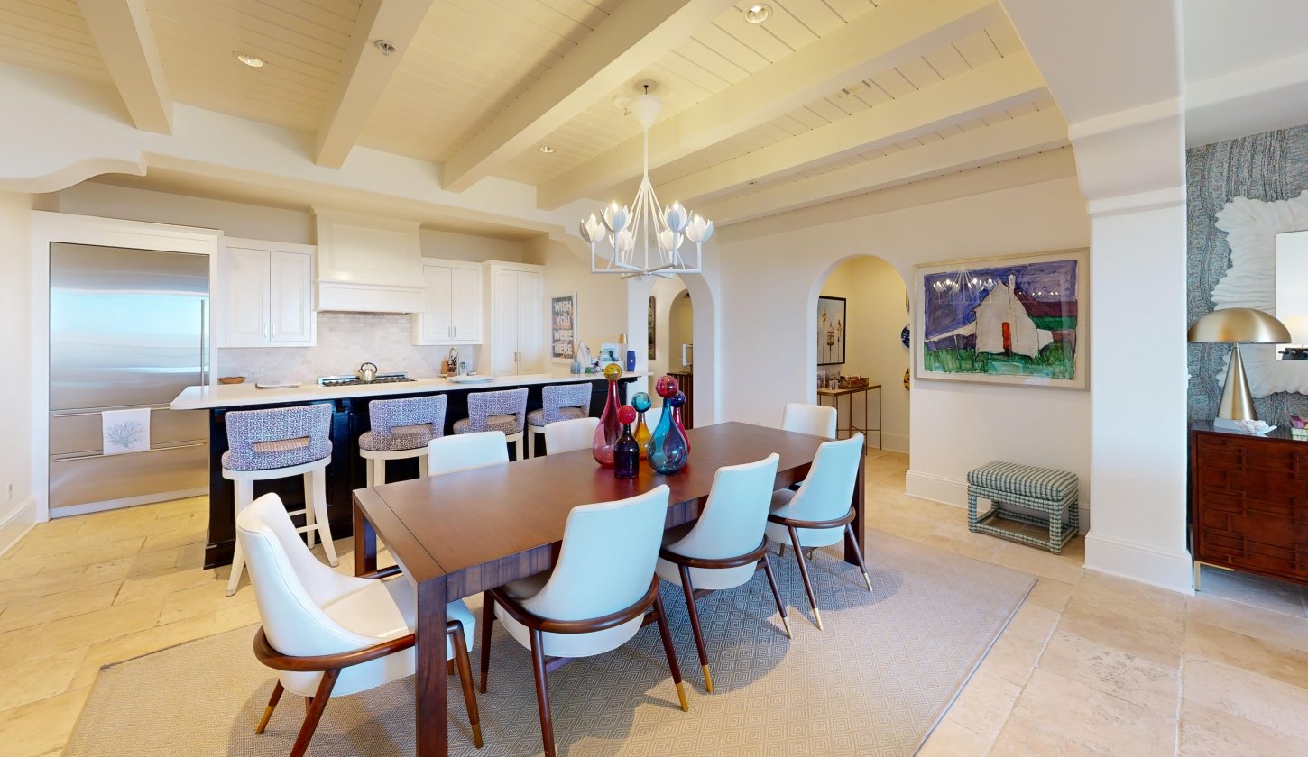 A long wooden dining table underneath a white chandelier with a kitchen in the background