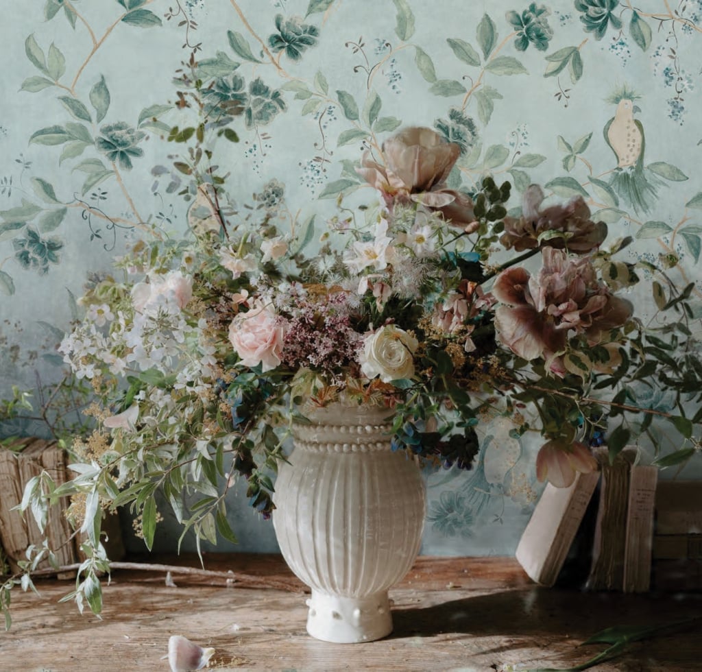 A white vase of flowers site in front of blue wallpaper