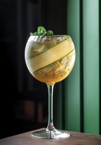 Elegant cocktail with ice, cucumber ribbon, and mint garnish served in a wine glass on a dark wooden table with a green vertical panel background.